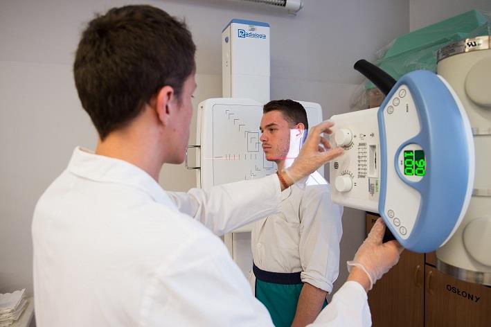 na zdjęciu widać dwóch uczniów którzy znajdują się przy szkolnym aparacie do robienia zdjęć rentgenowskich. Jeden uczeń ustawia aparat, drugi uczeń udaje pacjenta