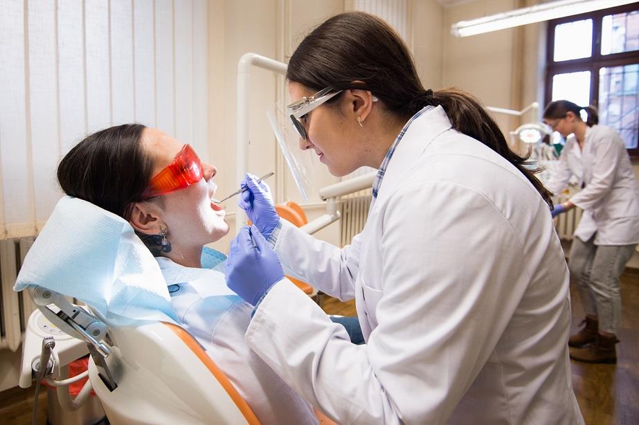  na fotografii znajdują się dwie uczennice uczące się wykonywania zabiegów stomatologicznych. Jedna uczennica trzyma narzędzia dentystyczne, druga uczennica siedzi na fotelu i udaje pacjenta