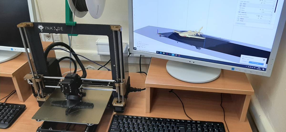 Fotografia przedstawia pracownię druku 3D, w której znajdują się komputer oraz drukarka 3D.