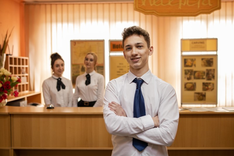 Na pierwszym planie stoi uczeń w lobby hotelowym, za nim dwie uczennice za ladą recepcji.