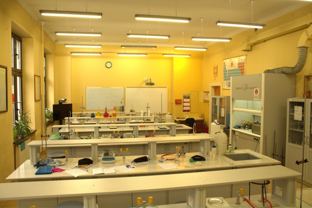 Fotografia przedstawia szkolną pracownię chemiczną. W pracowni znajduje się digestorium oraz kilkanaście stanowisk, przy których uczniowie mogą przeprowadzać eksperymenty chemiczne przy użyciu probówek i szalek.